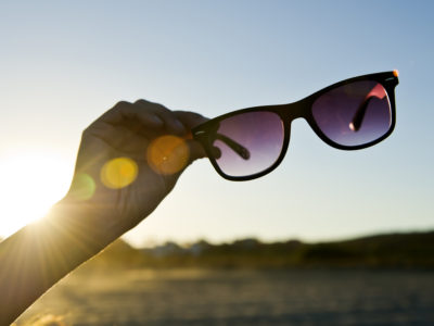 Hand Holding Black Sunglasses at sunset in front of vibrant blue sky. The Sun iluminates de scene with the golden light of dawning. Backlit