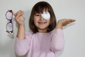 Young girl smiles with an eye patch on and holds her glasses in one hand