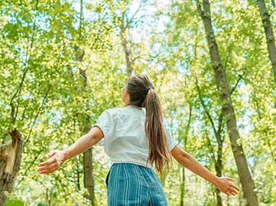 Free woman breathing clean air in nature forest. Happy girl from the back with open arms in happiness. Fresh outdoor woods, wellness healthy lifestyle concept.