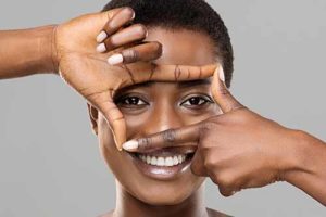 Blepharoplasty concept. Beautiful smiling black woman holding frame from fingers near her eyes, enjoying result of plastic surgery, gray background