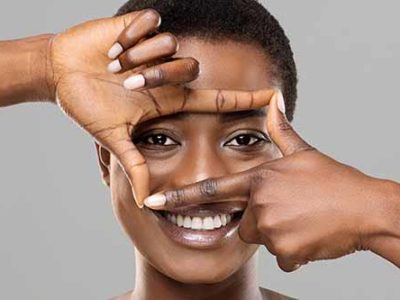 Blepharoplasty concept. Beautiful smiling black woman holding frame from fingers near her eyes, enjoying result of plastic surgery, gray background