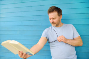 Portrait of mature man with big black eye glasses trying to read book but having difficulties seeing text because of vision problems