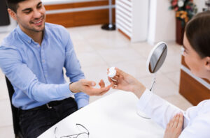 Vision Correction Concept. Female doctor optometrist giving plastic container with contacts lens to smiling male patient customer sitting at table. Healthcare and consultation at the optics store